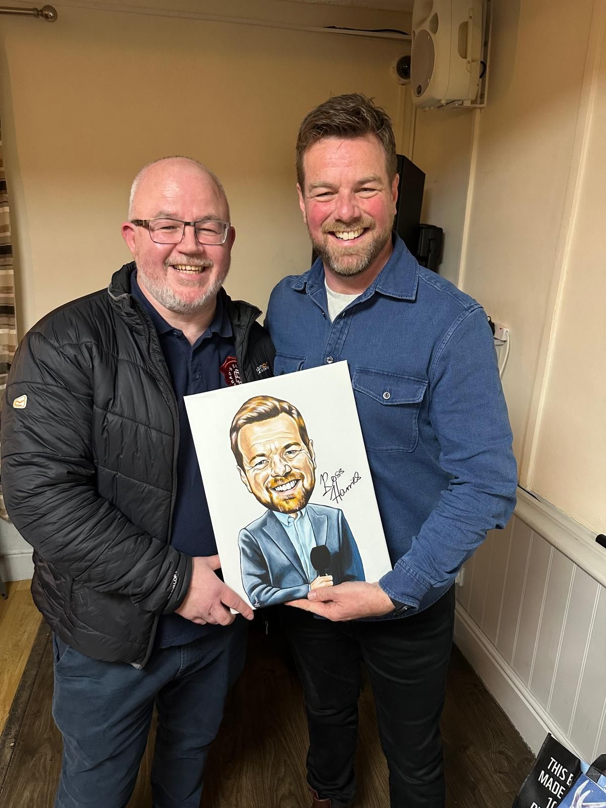 Two smiling men hold a caricature portrait in a hallway