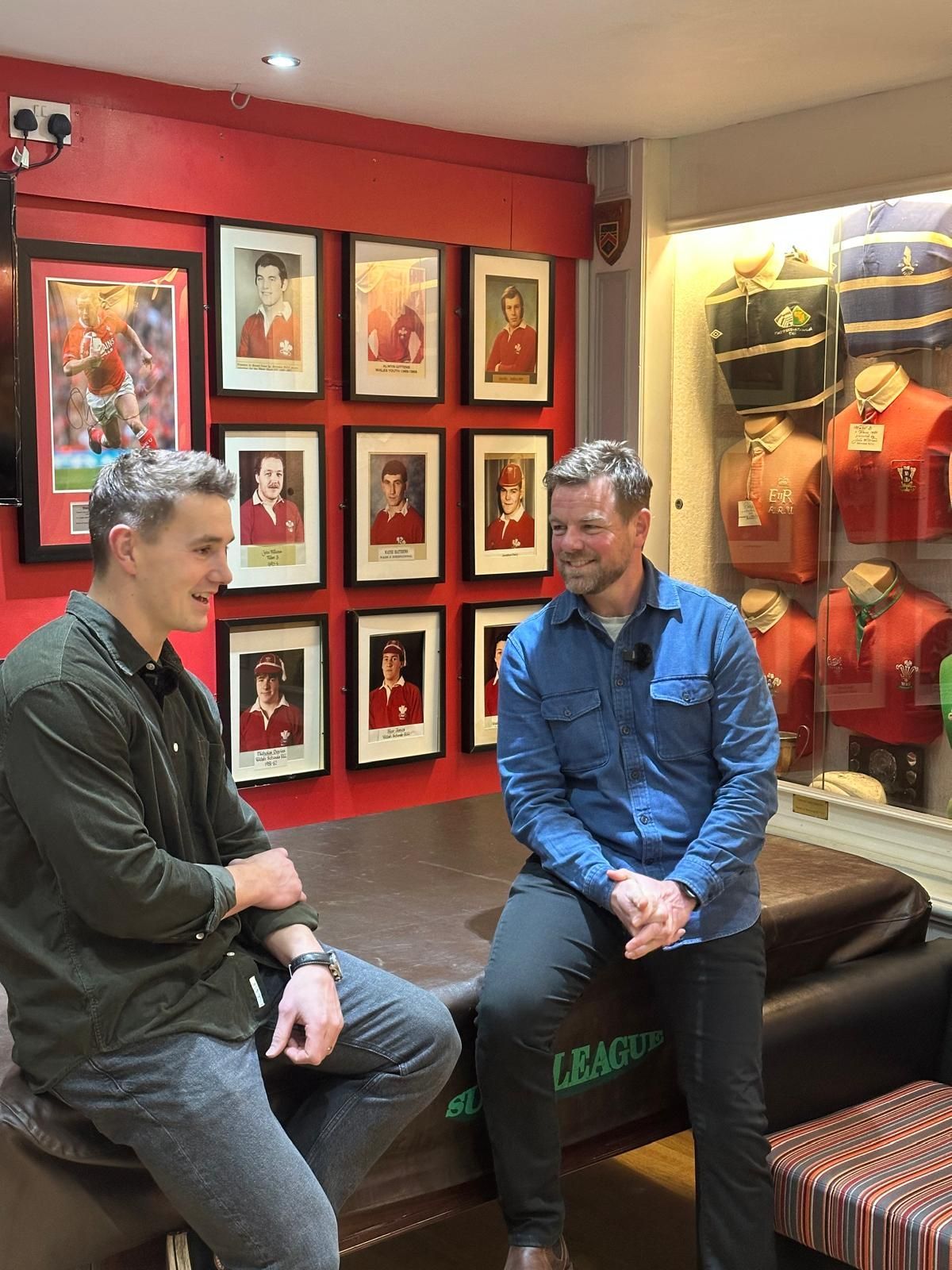 Two people sit and talk in a red-walled room with framed portraits and sports jerseys.