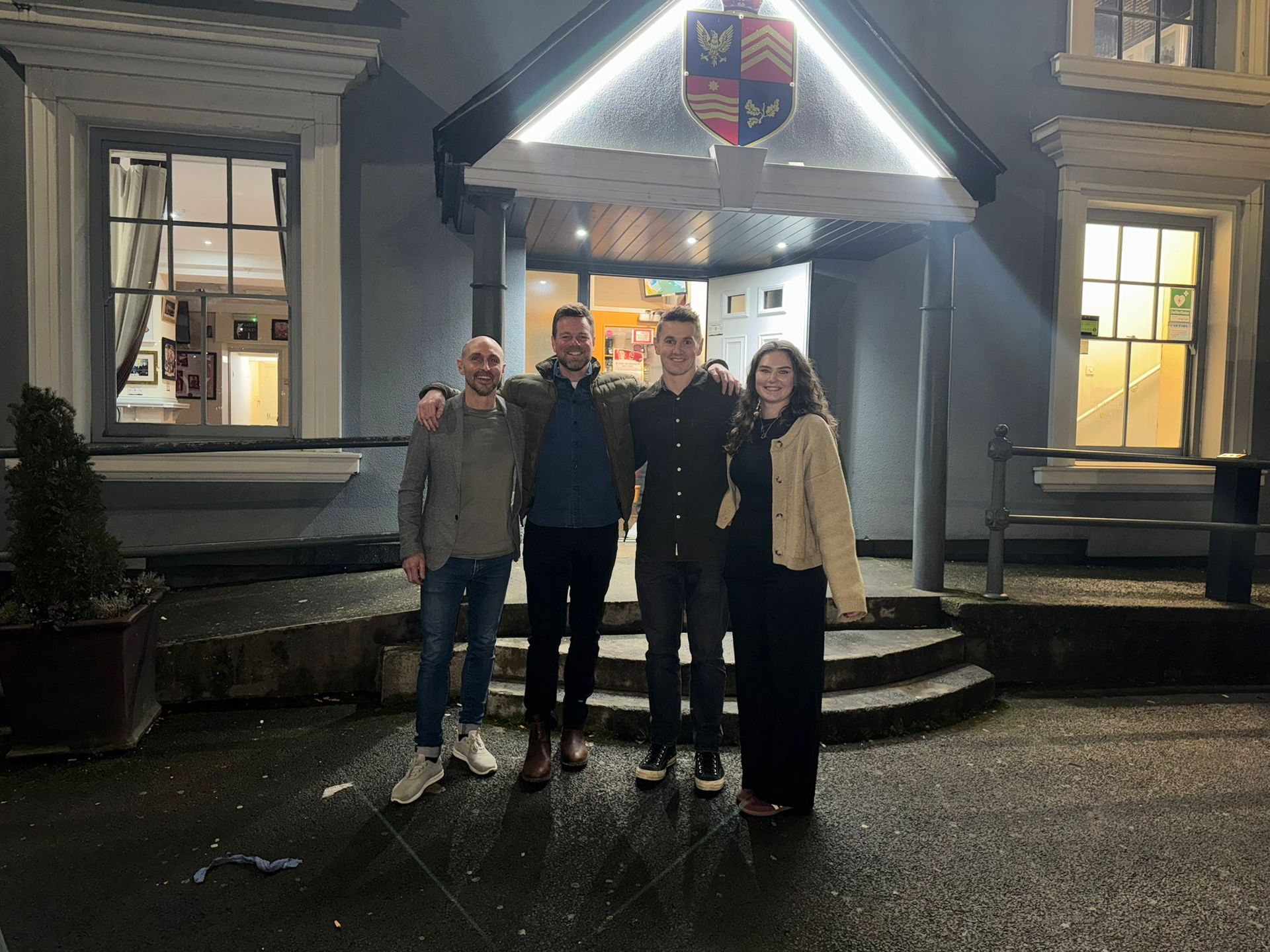 Four people posing outside a lit building entrance at night, with a crest above the doorway.