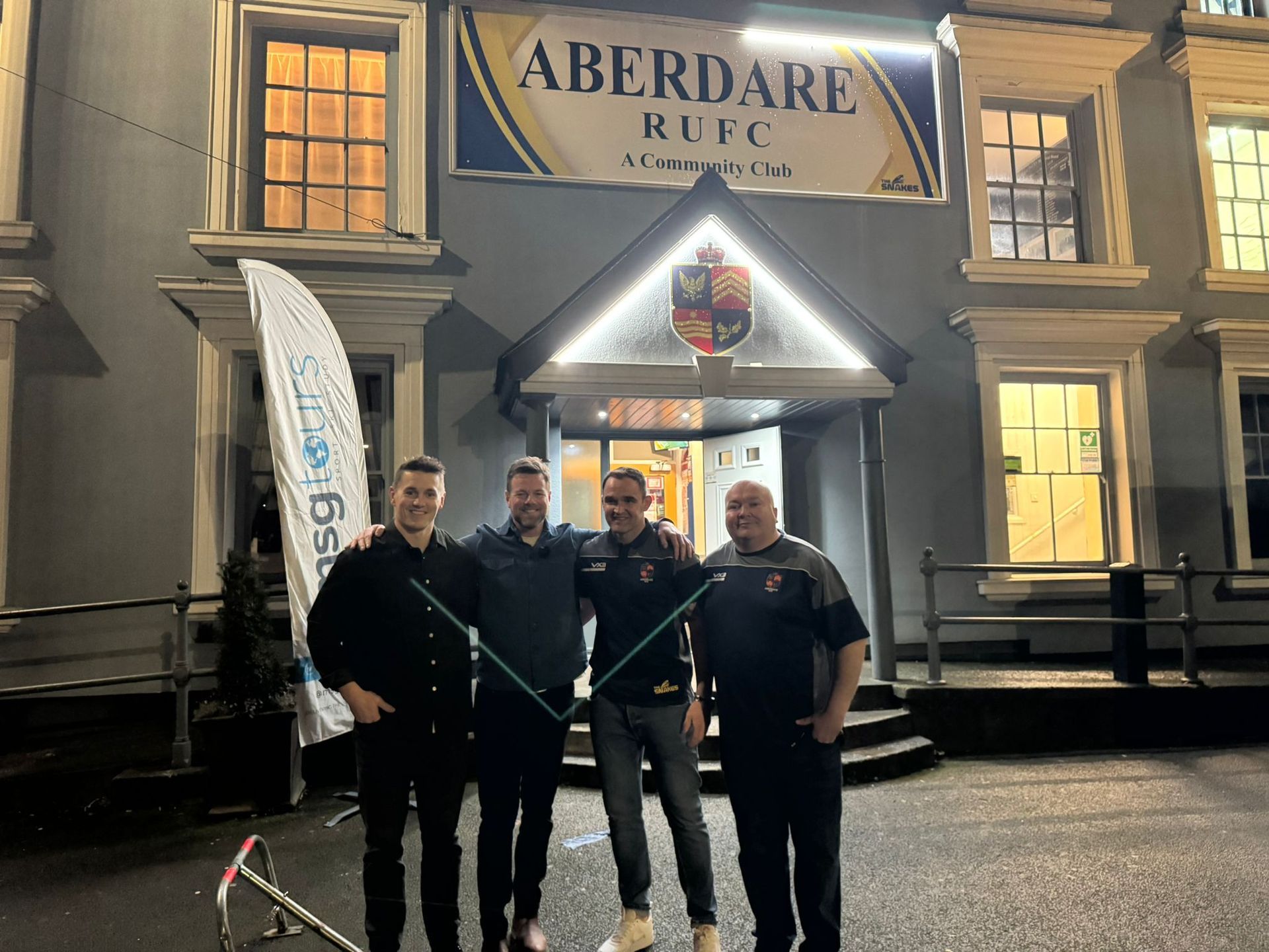 Four men standing outside Aberdare RFC at night, with a lit entrance and rugby club sign behind them.