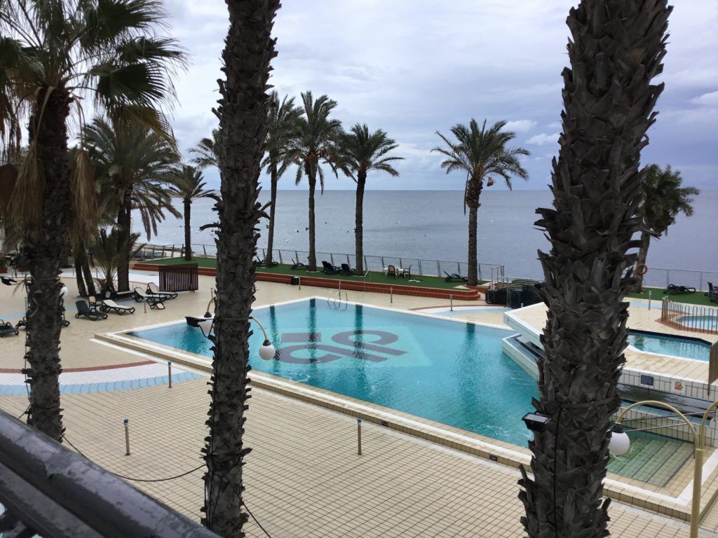Palm-lined resort pool with lounge chairs overlooking a calm ocean under a cloudy sky