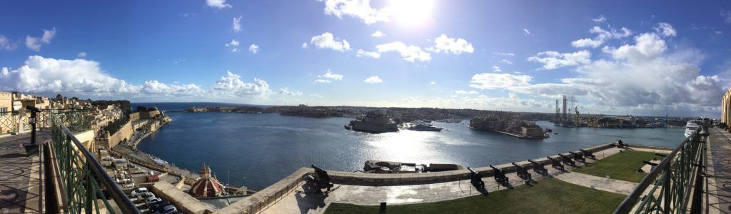 Panoramic harbor view with blue water, boats, city skyline, and sunny sky with clouds