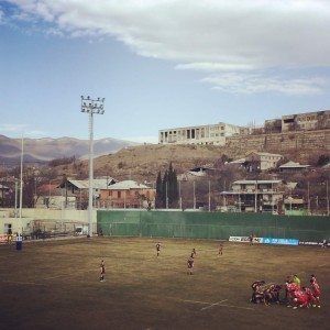 Soccer match on a dusty field with players, a green fence, floodlight, and hillside buildings in the background