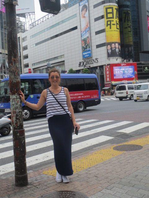 Woman standing on a city sidewalk at a crosswalk, holding a pole with buses and buildings in the background.
