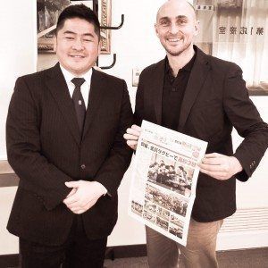 Two men in suits posing indoors, one holding a newspaper, with Japanese text on the wall.