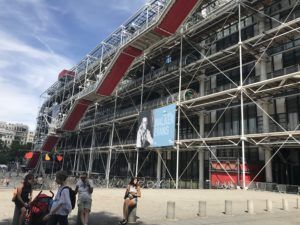 Modern building with exposed steel scaffolding and red stairways, people walking in front outside.