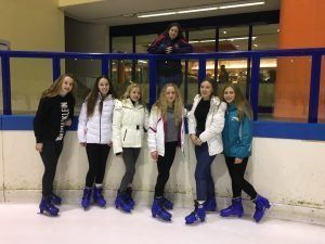 Six people in winter gear stand on an indoor ice rink, wearing blue skate covers and posing by the boards.