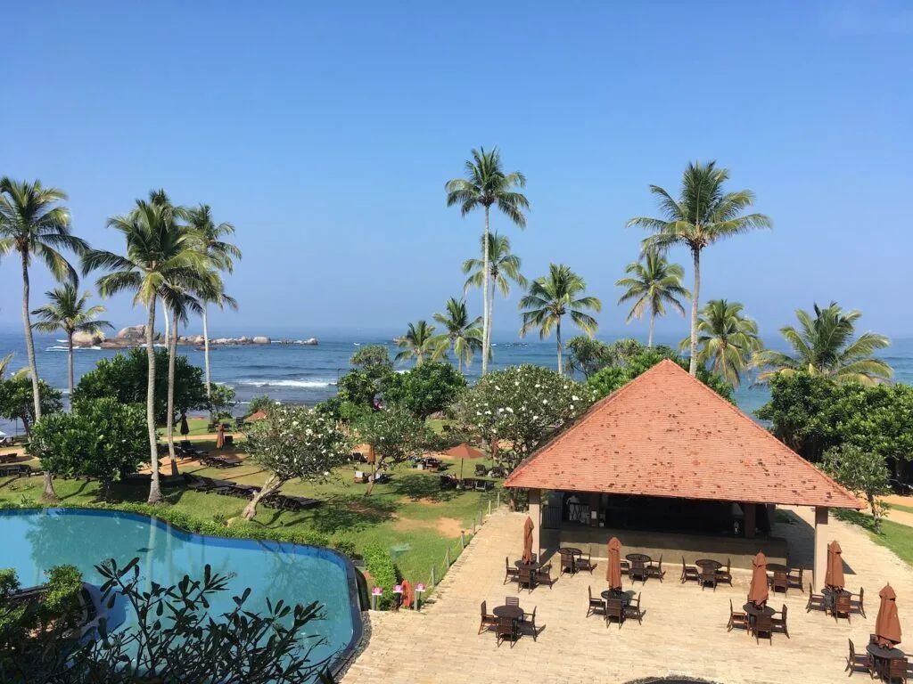 Tropical resort with palm trees, ocean view, pool, and a red-roofed patio with tables and chairs