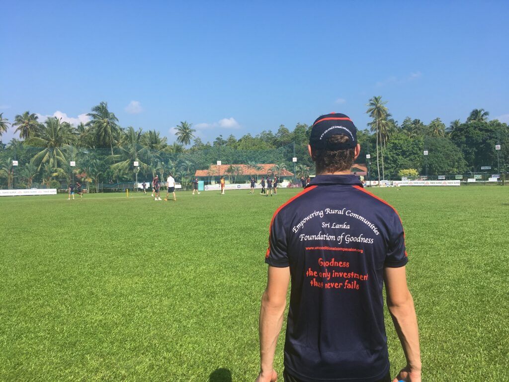 Person in a black jersey stands on a grassy field, facing distant players and trees under a clear blue sky.