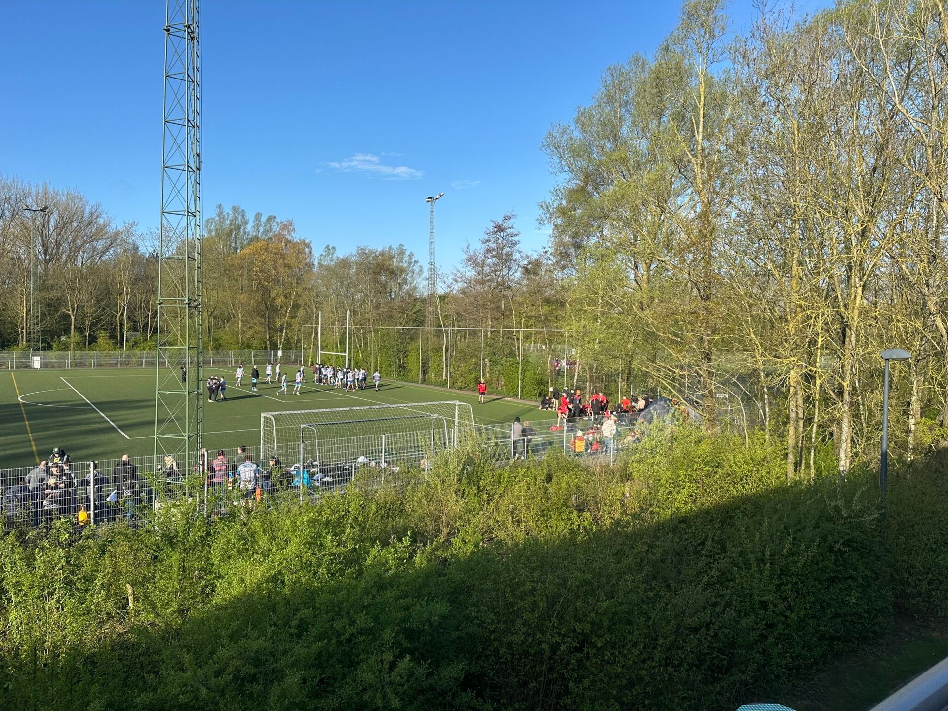 People gathered near a fenced sports field beside trees on a sunny day