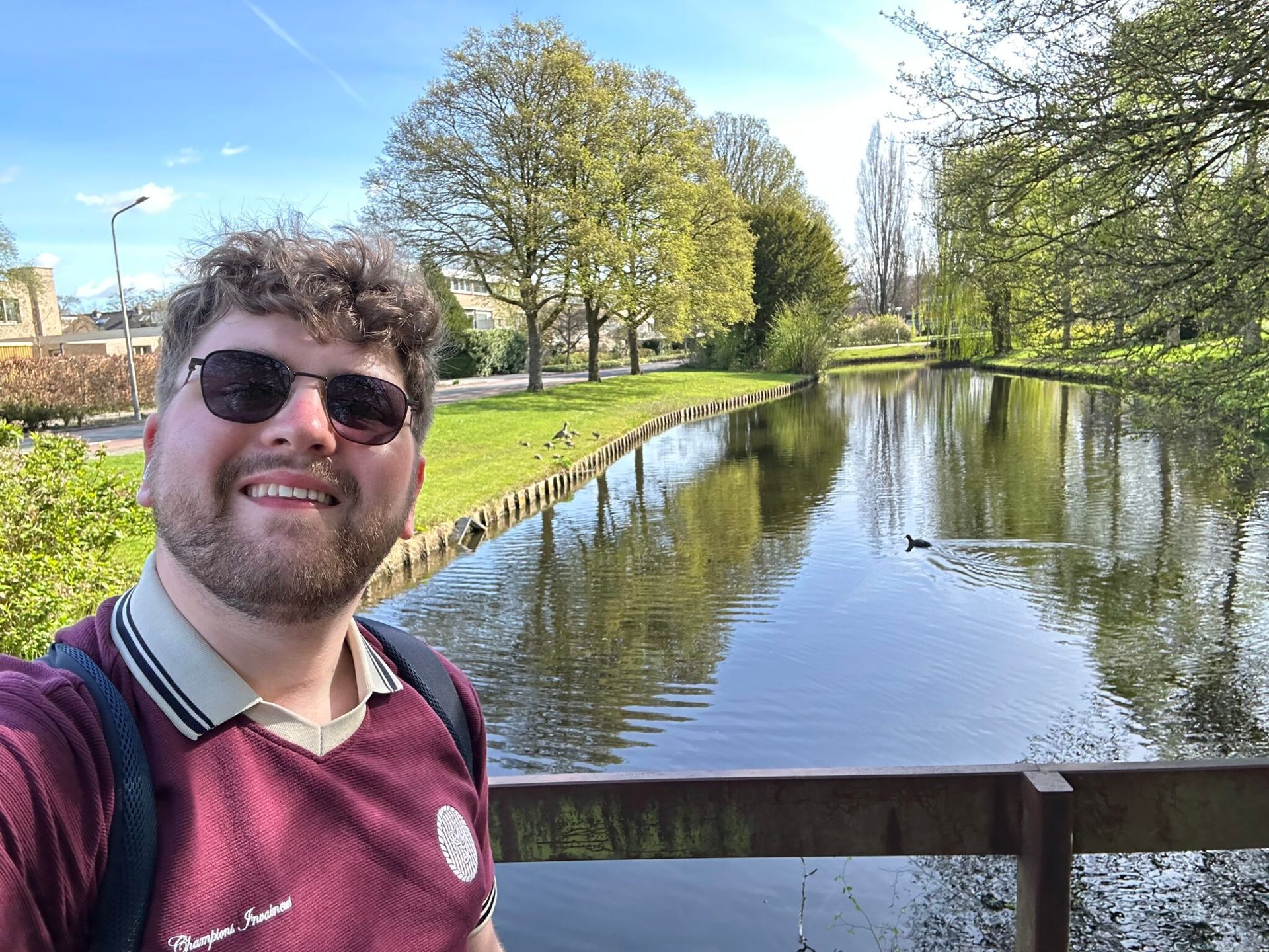 Smiling person in sunglasses by a canal with trees and blue sky reflected in the water