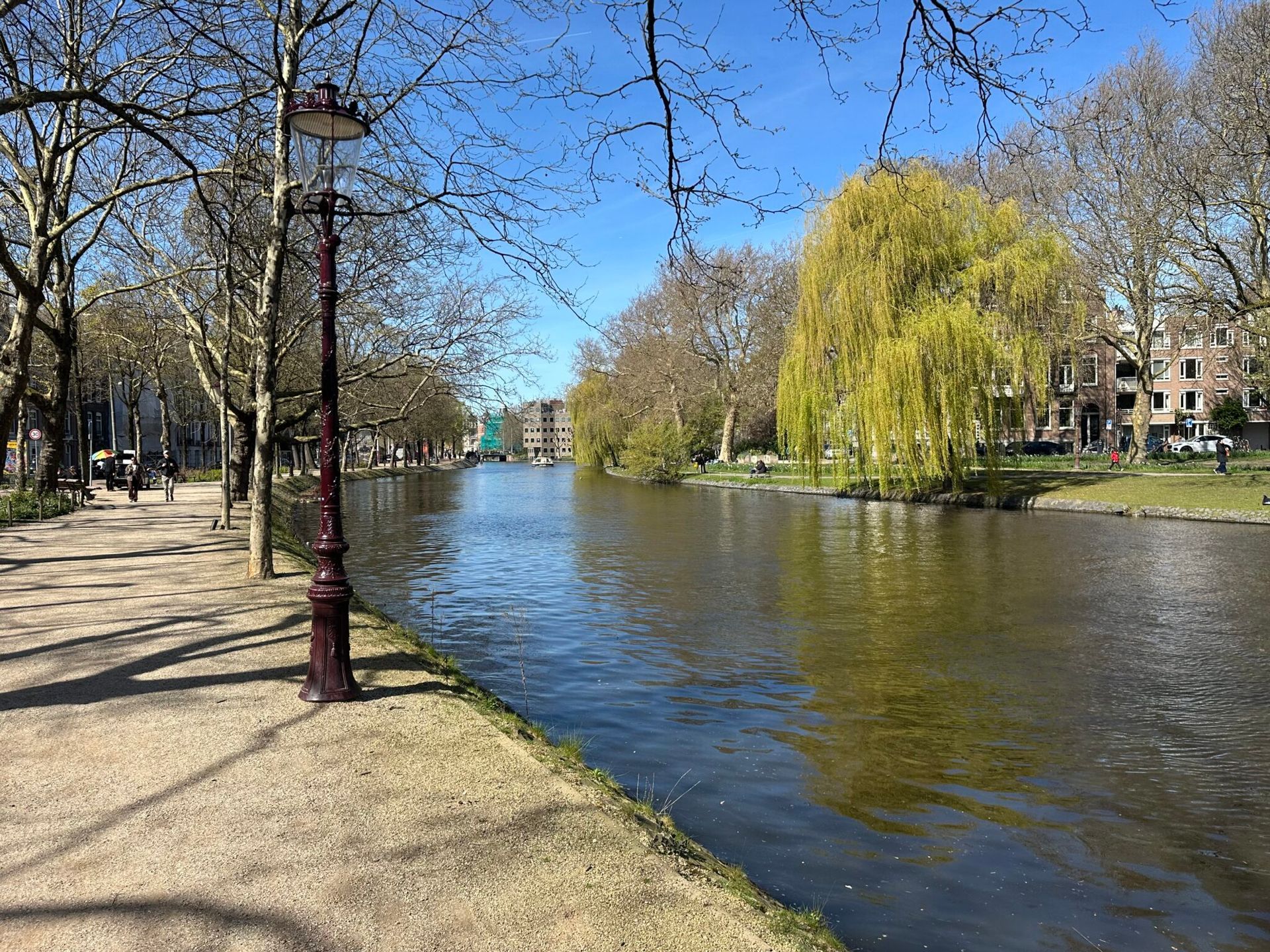 Canal lined with trees and lampposts beside a gravel path on a sunny day