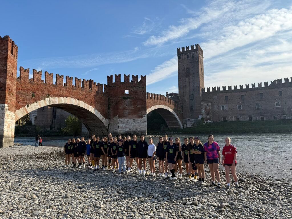 Group posing on a pebble beach before a brick medieval castle and arched bridge under a blue sky
