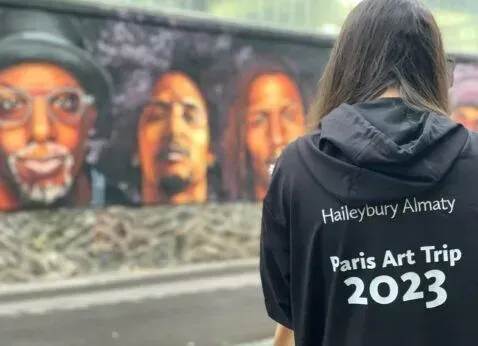 Person in black hoodie facing a mural; back text reads “Haileybury Almaty” and “Paris Art Trip 2023.”