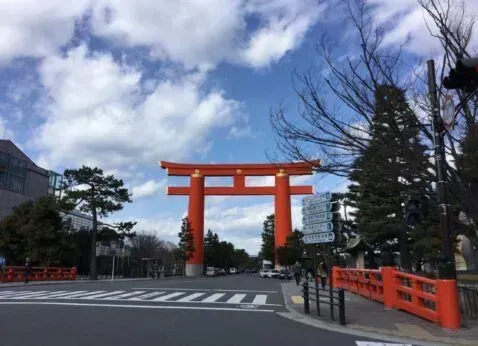 Red torii gate over a road, with trees, buildings, and a cloudy blue sky.