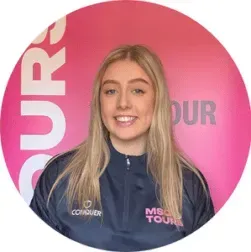 Person in navy jacket smiling before a pink circular “TOUR” backdrop