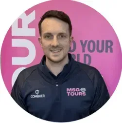 Person in a navy MSQ Tours jacket smiling in front of a pink branded backdrop.