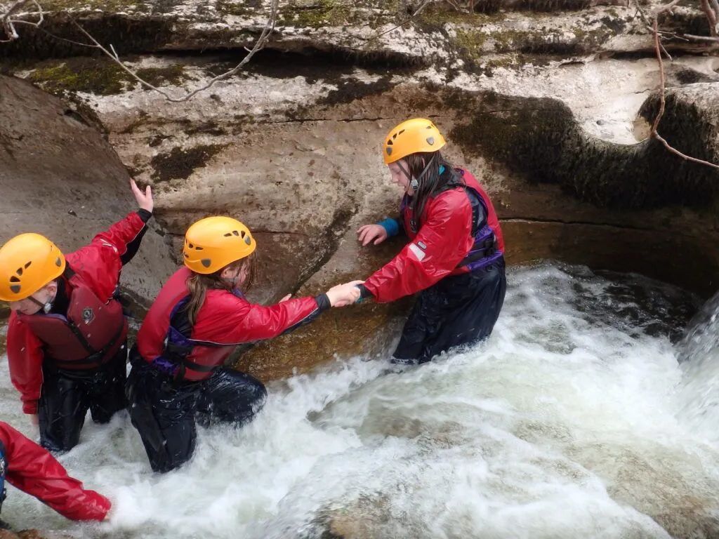 Three people in yellow helmets and red jackets crossing a rocky stream in whitewater￼