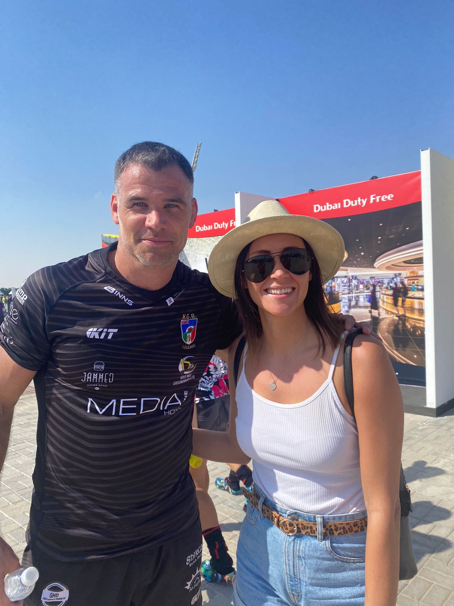 Two people smiling outdoors, one in a black jersey and one in a straw hat and sunglasses, at a sunny event booth