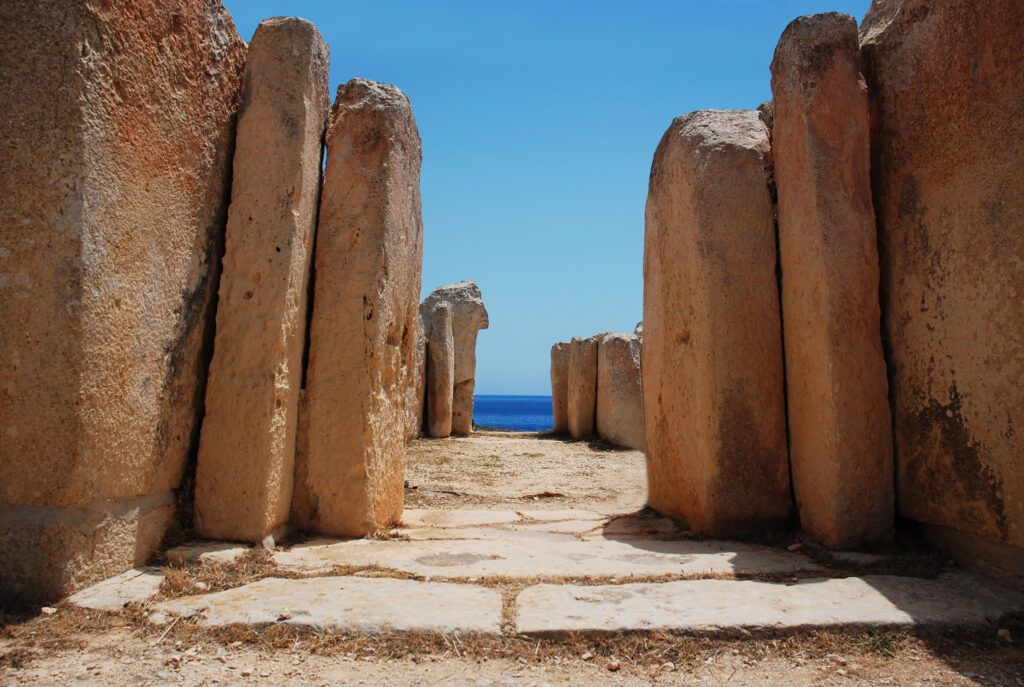 Ancient stone pillars lining a sandy path leading to the blue sea under a clear sky