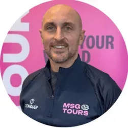 Smiling bald man in navy MSG Tours jacket standing before a pink backdrop