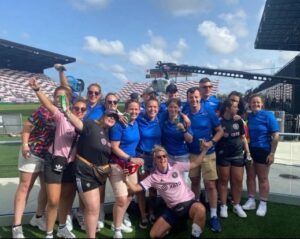 Group of people in blue shirts and athletic wear posing on a sports field, cheering and smiling.