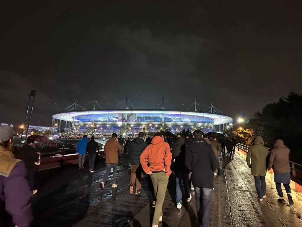 Crowd walking toward a brightly lit stadium at night in the rain