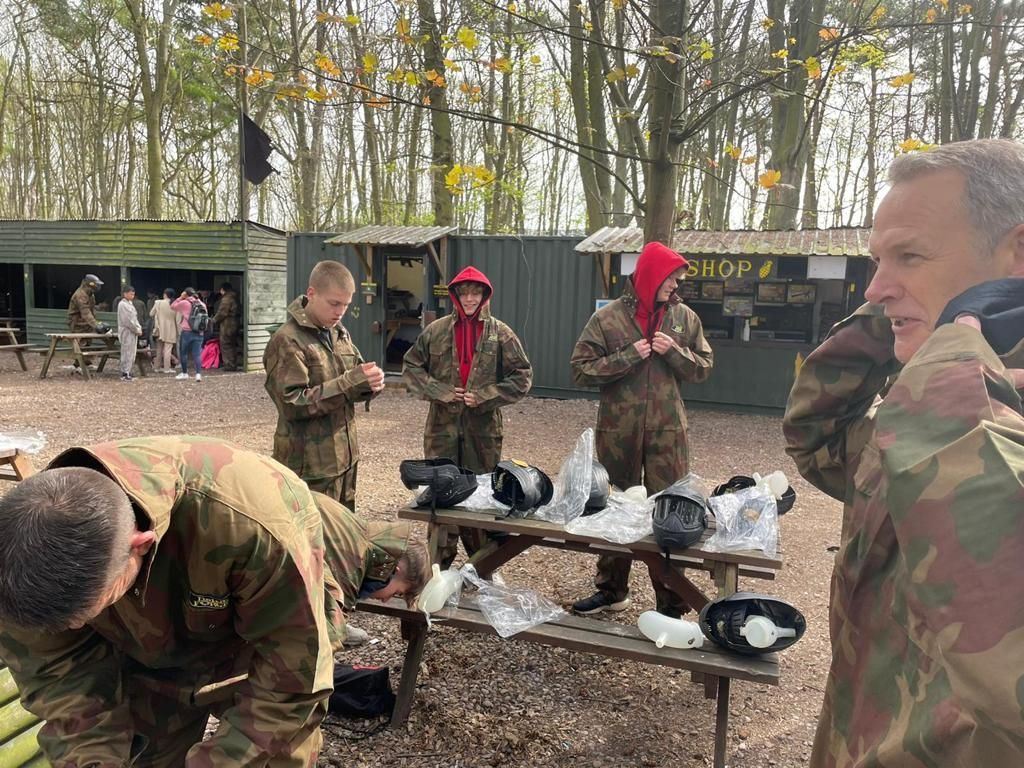 Camouflage-clad people gathering and eating outdoors at a wooded campsite with tents and tables