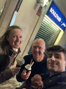 Three people smiling with ice cream cones outside a shop at night under a blue sign