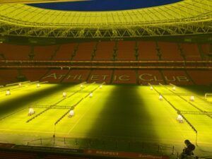 Soccer match on a sunlit green field in a large stadium with red stands and a yellow roof.
