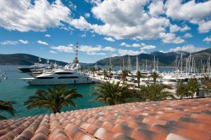 Yachts docked in a marina with mountains, palm trees, and a red-tiled roof in the foreground under a blue sky.