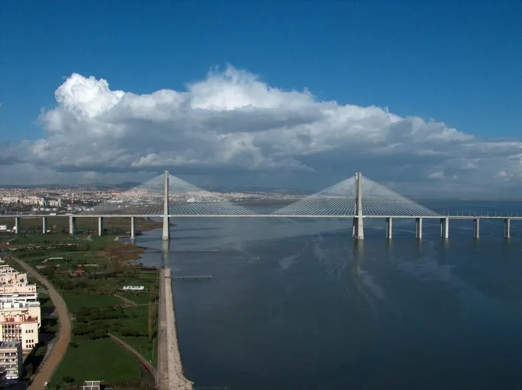 Cable-stayed bridge spanning a wide river under a cloudy blue sky, with city buildings at left