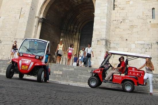 Two red tuk-tuks parked by a stone building with a large arched entrance and people on the steps
