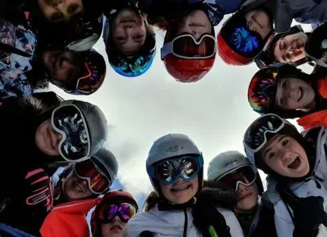 Snowboarders in helmets and goggles circle the camera, smiling up against a bright sky