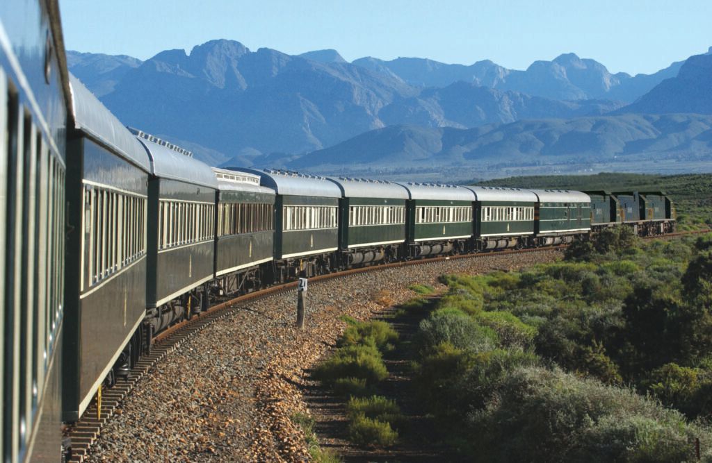 Curving passenger train in a mountain landscape with blue peaks and dry, rocky terrain