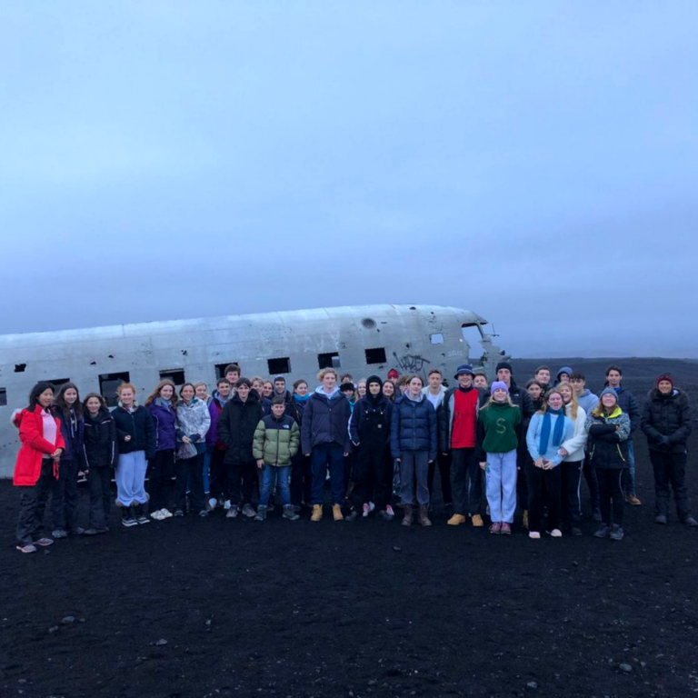 Group of people posing in front of a large airplane wreck on a dark beach under a cloudy blue sky