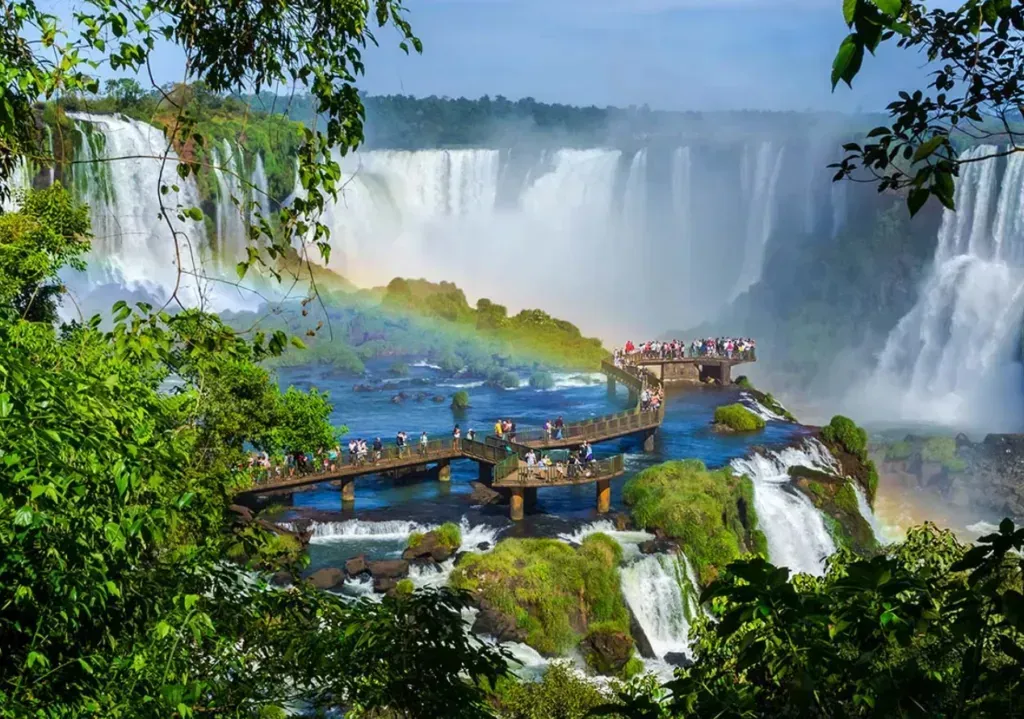 Tourists on a walkway overlooking a lush, misty waterfall with rainbows at Iguazu Falls