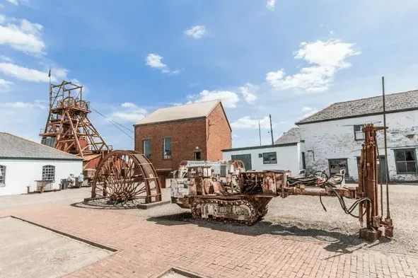 Rusty industrial mining machinery displayed in a courtyard with brick and white buildings under a blue sky