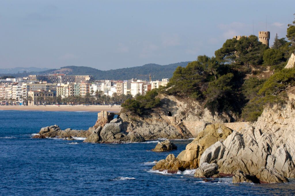 Rocky coastline beside a blue sea, with a distant coastal town and hills under a clear sky.
