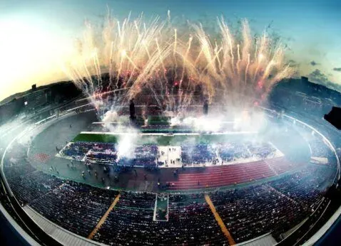 Fireworks over a packed stadium during a nighttime event, with bright lights and smoke filling the sky