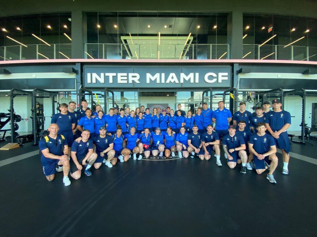 Large group posing in front of an INTER MIAMI CF sign, wearing blue shirts in a gym-like facility