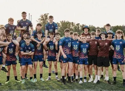 Rugby team posing on a grassy field, wearing blue and maroon jerseys at sunset