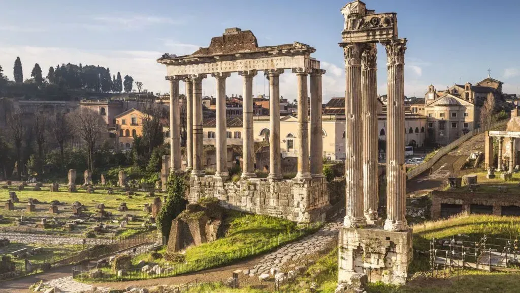 Roman ruins with tall stone columns and arches overlooking a grassy ancient cityscape under a clear sky