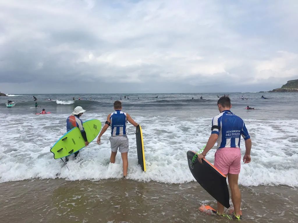 Three surfers wade into the ocean with boards at a cloudy beach.
