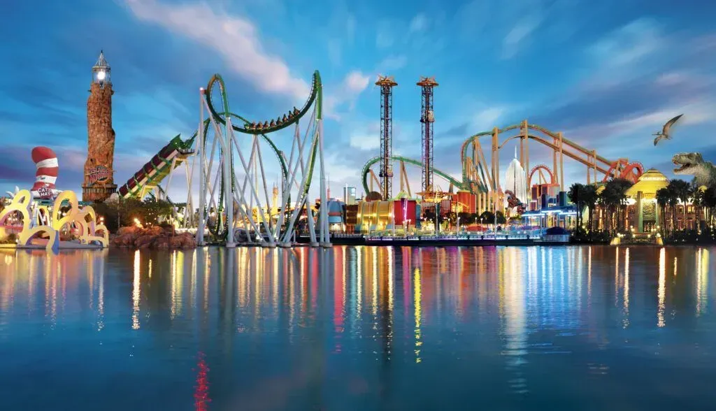 Colorful amusement park rides reflected on calm water at dusk, with a bright blue sky.