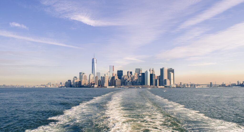 Skyline of Lower Manhattan viewed from a boat wake on blue water under a partly cloudy sky