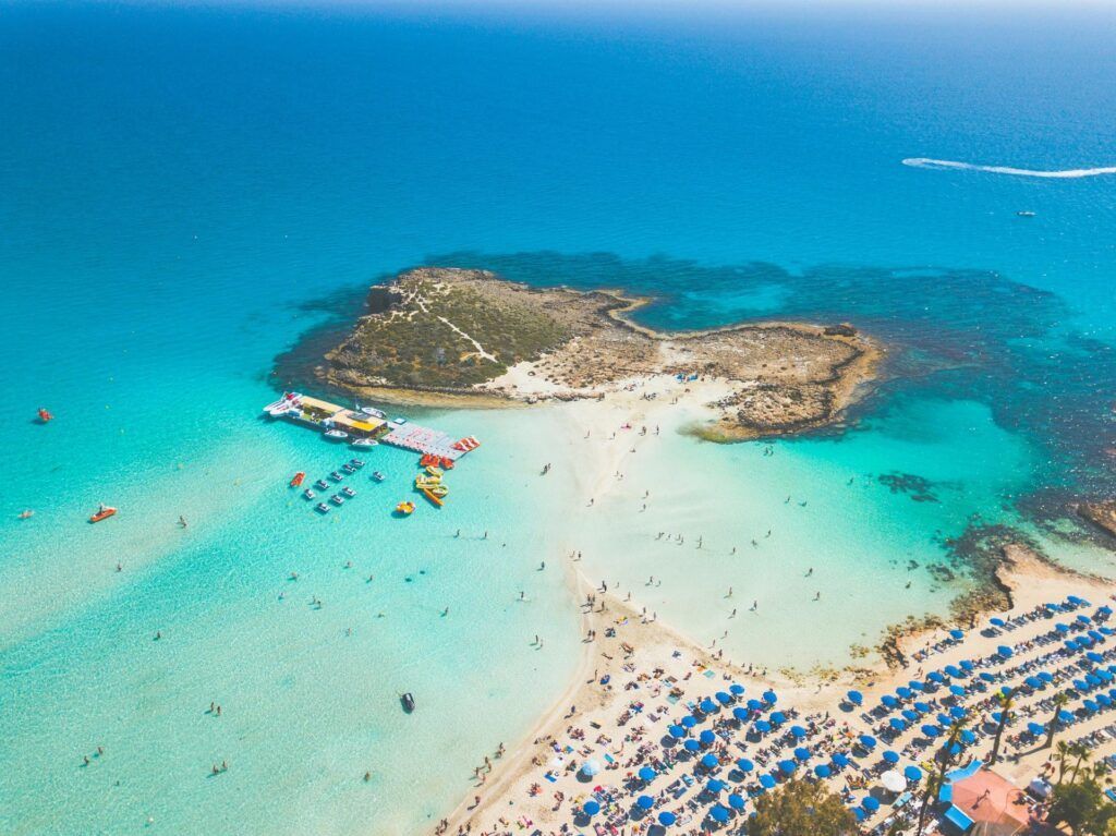 Aerial view of a turquoise beach with a rocky island, swimmers, and rows of blue umbrellas on the shore