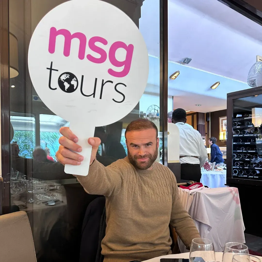 Man holding a “msg tours” sign in a restaurant or café, seated by a window