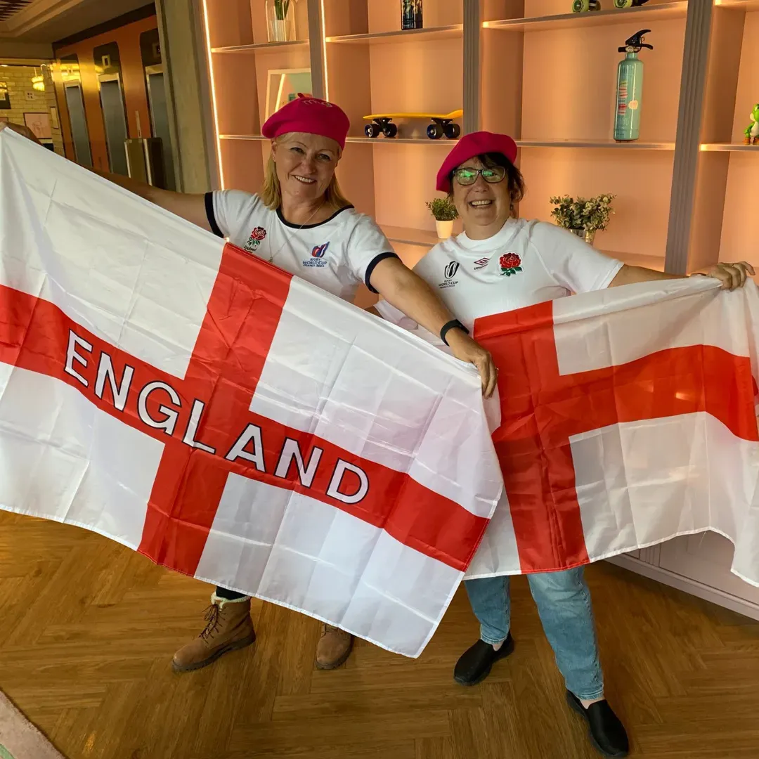 Two people holding England flags, wearing red hats, standing indoors and smiling.