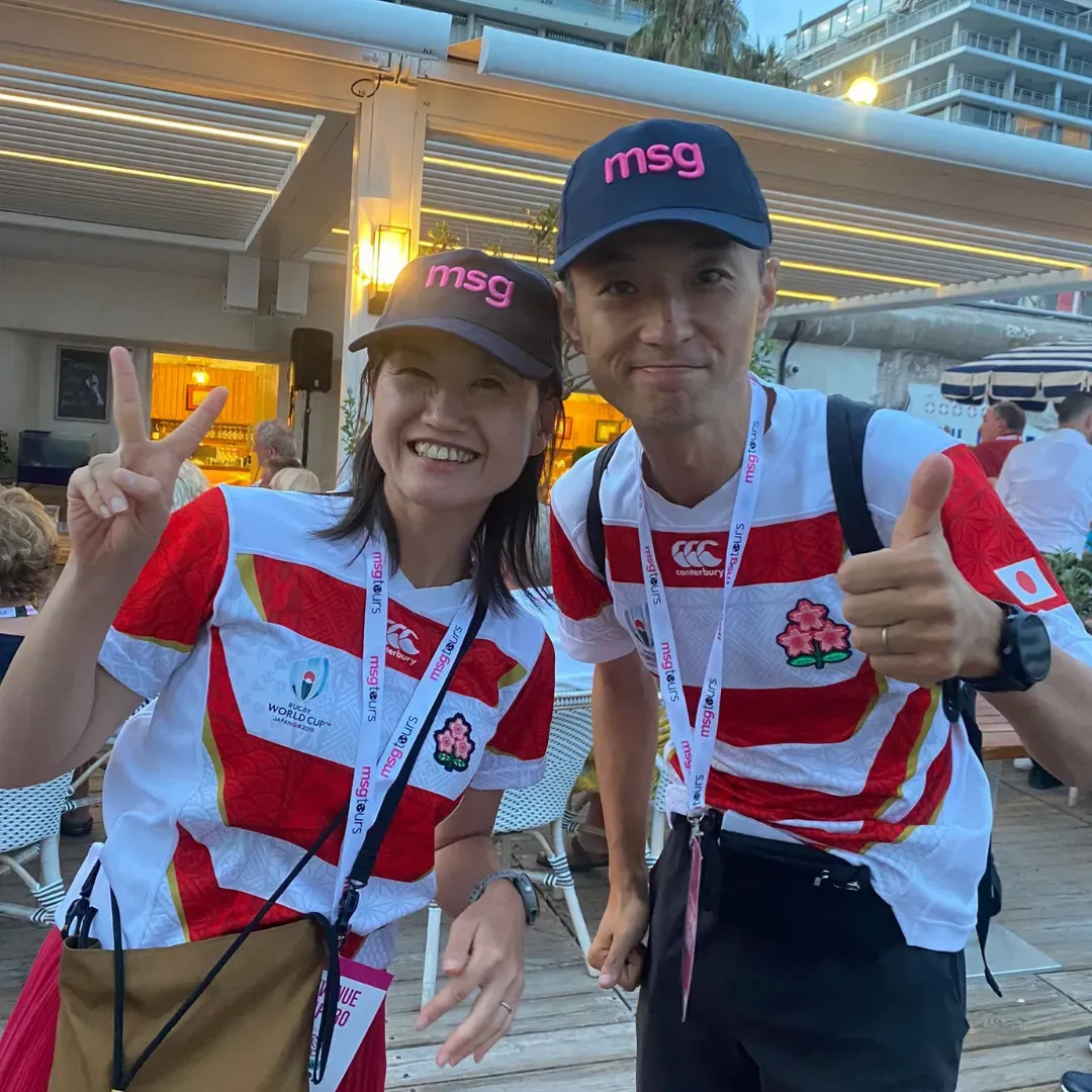 Two cyclists in red-and-white jerseys and black caps smiling and giving thumbs up outdoors at a transit stop